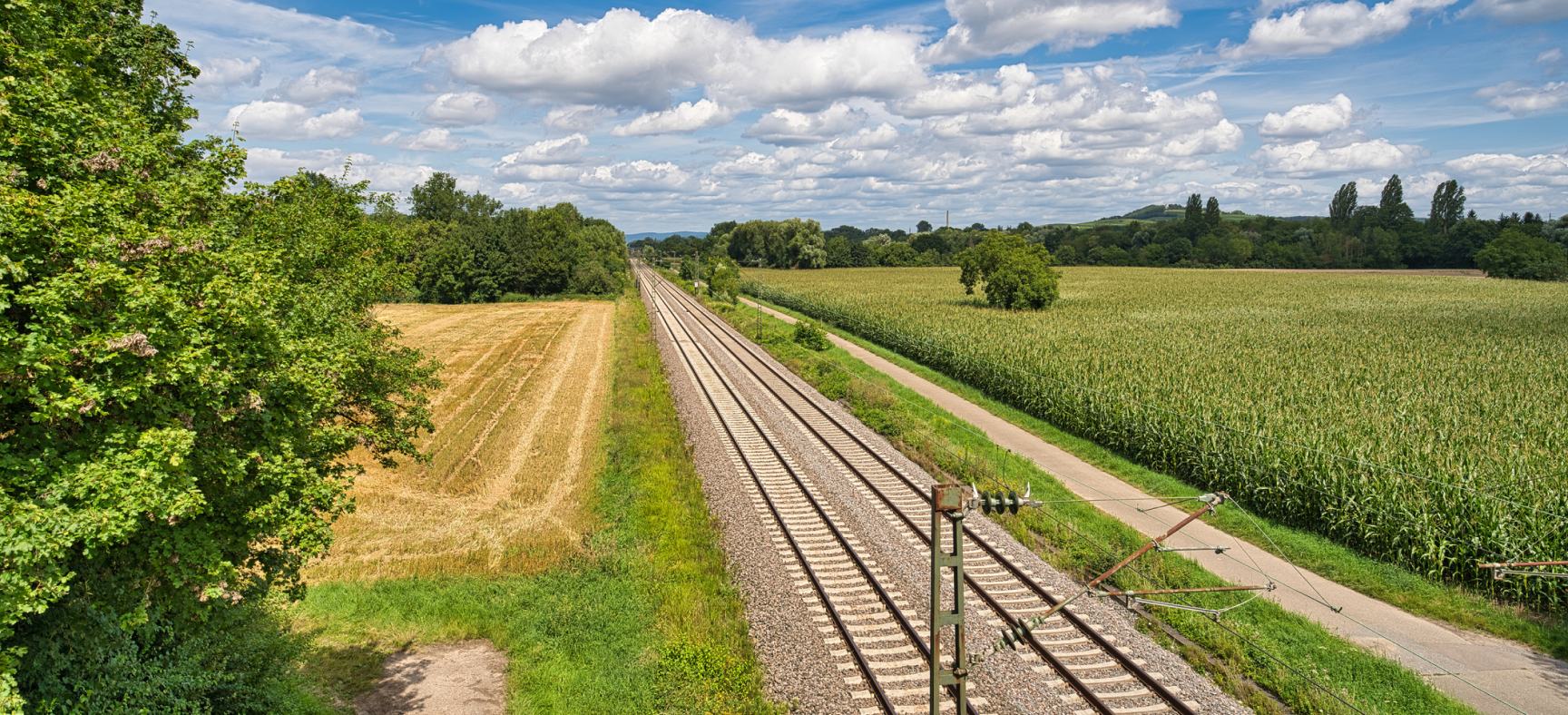 Wide angle view of train tracks with overhead electrical wires passing between fields into the horizon.