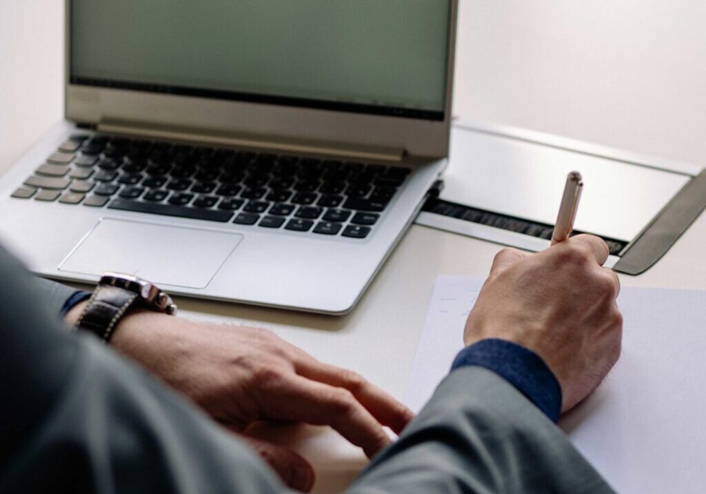 Une personne écrit avec un stylo et du papier à un bureau avec un ordinateur portable à côté d'elle.